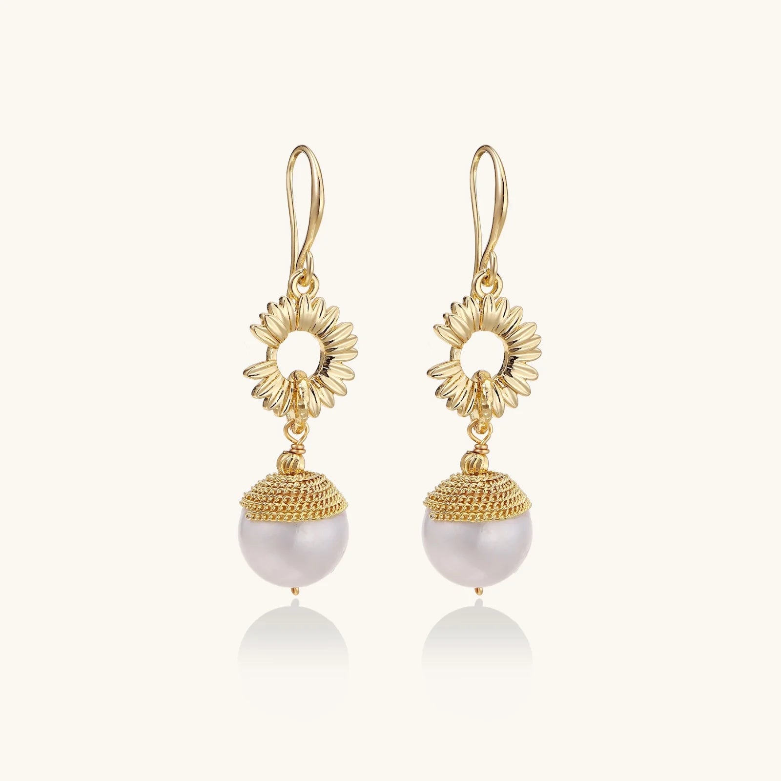 Gold sunburst charm earrings with baroque pearl drops on French hooks against cream background