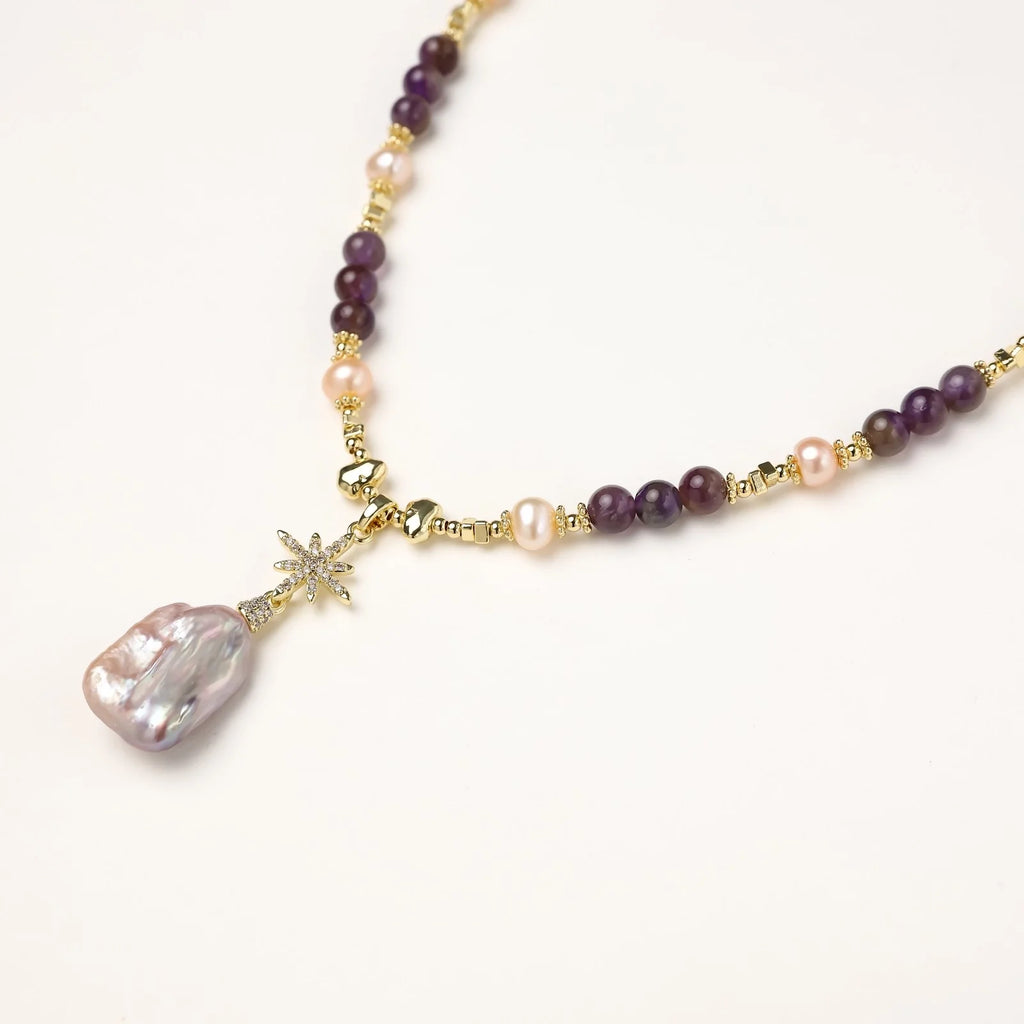 Gold necklace with iridescent baroque pearl pendant, purple amethyst beads, and crystal star charm on cream background